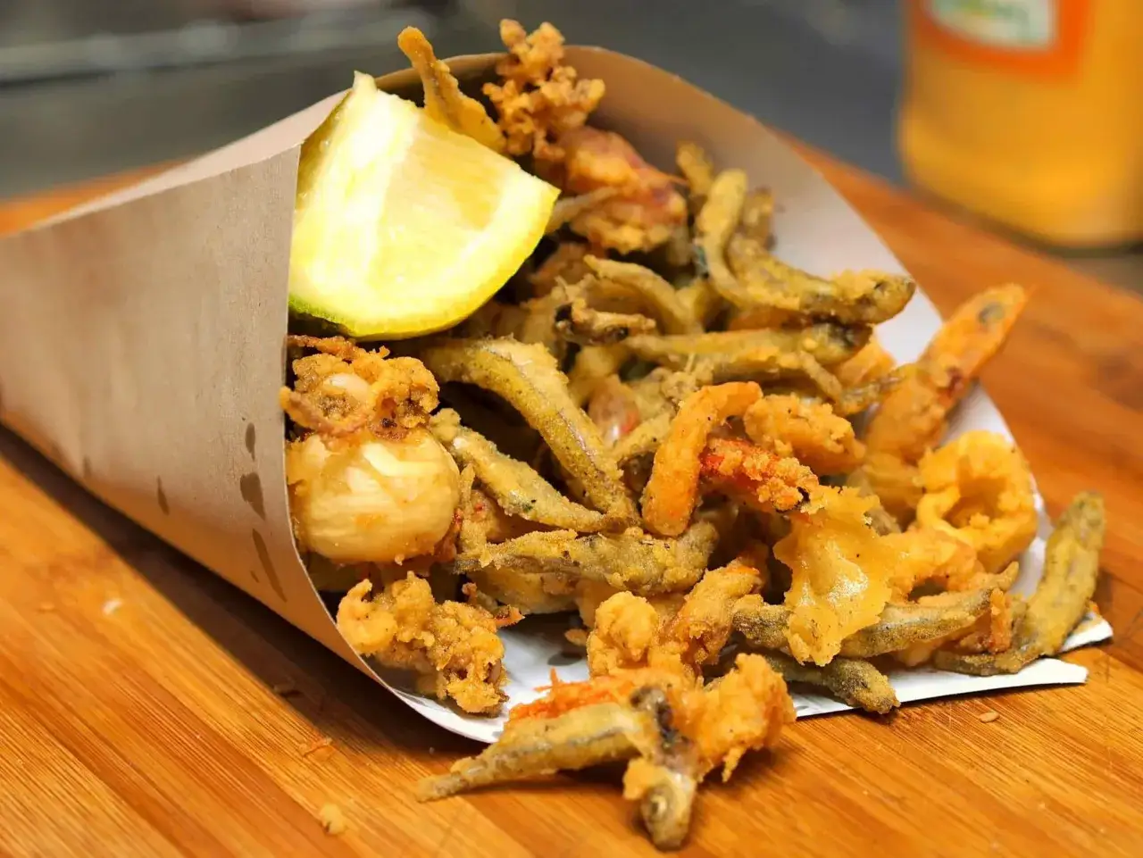 Un cono di carta marrone traboccante di una deliziosa frittura di pesce, con calamari, gamberi e pesciolini, guarnito con uno spicchio di limone.