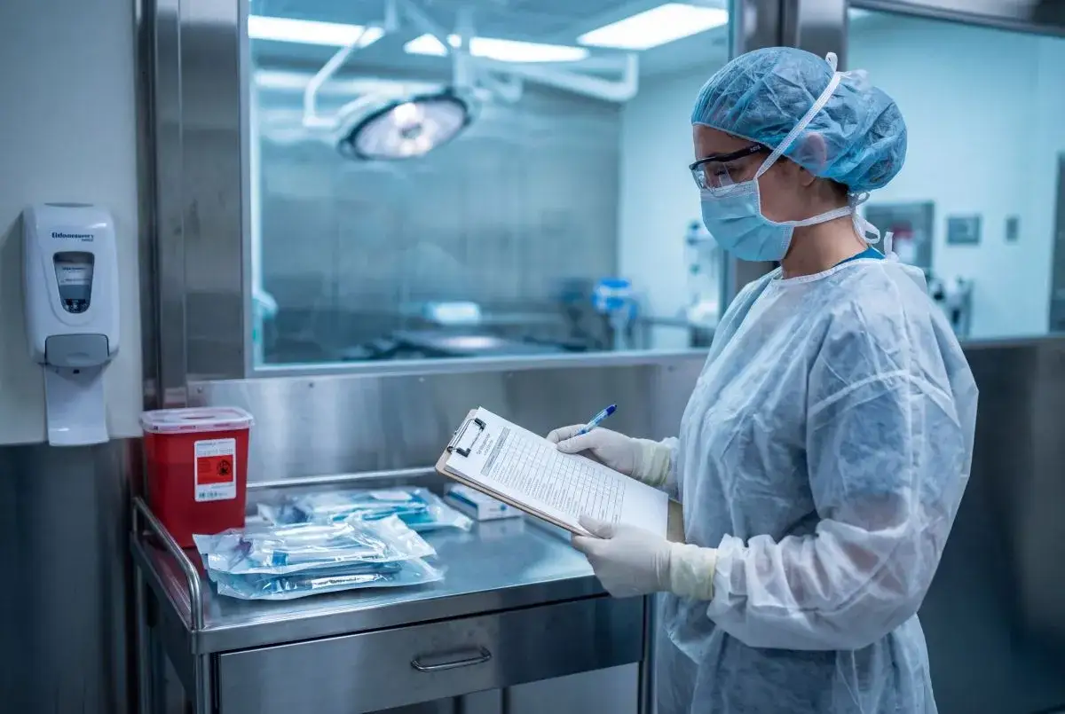 Pielęgniarka epidemiologiczna w fartuchu i masce notuje dane na kartce. W tle widać sterylne pomieszczenie.