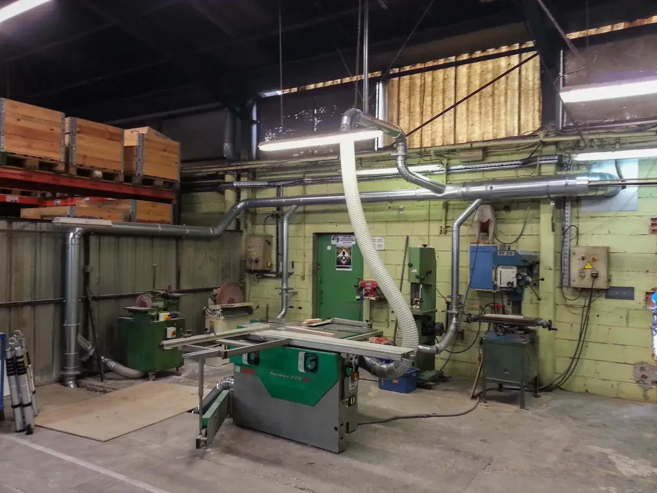 Vue d'un atelier de menuiserie bois avec machines, tuyaux d'aspiration et palettes de bois empil&eacute;es.