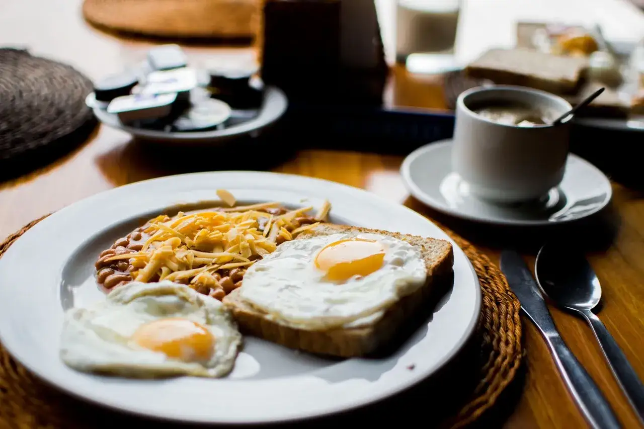 Un petit déjeuner sportif : œufs au plat, haricots et fromage râpé sur une assiette blanche, avec une tasse de café à côté.