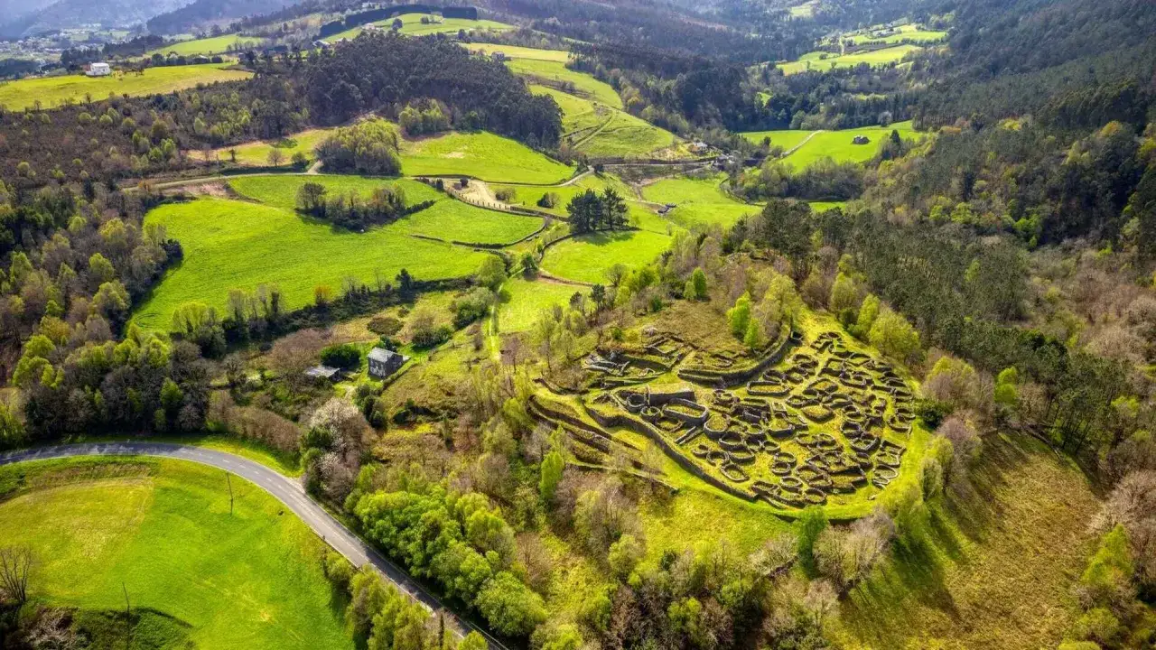 Vista aérea de un poblado celta en ruinas, con sus muros circulares de piedra integrados en un paisaje verde y ondulado de colinas y bosques.