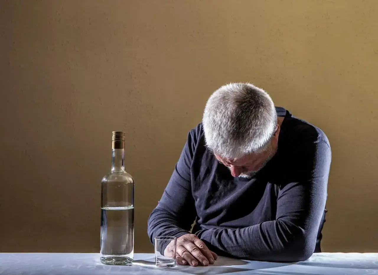 Mężczyzna z siwymi włosami pochyla głowę nad stołem obok butelki alkoholu i pustej szklanki. To obraz, który może ilustrować asertywne odmawianie picia alkoholu przykłady.