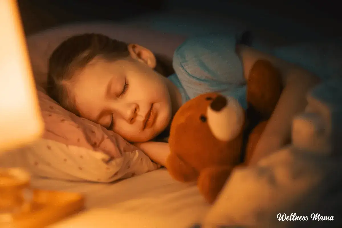 Dziecko śpi z misiem, przytulone do poduszki. Delikatne światło lampki nocnej rozprasza lęk przed ciemnością.