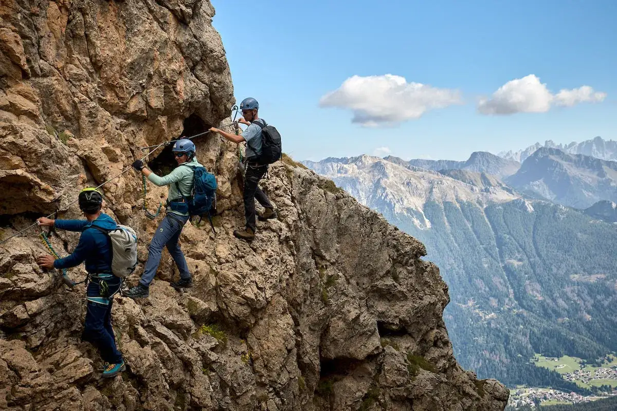 Trzech turystów pokonuje via ferrata w Dolomitach, zabezpieczeni linami na skalnej ścianie. W tle rozciąga się majestatyczny górski krajobraz.