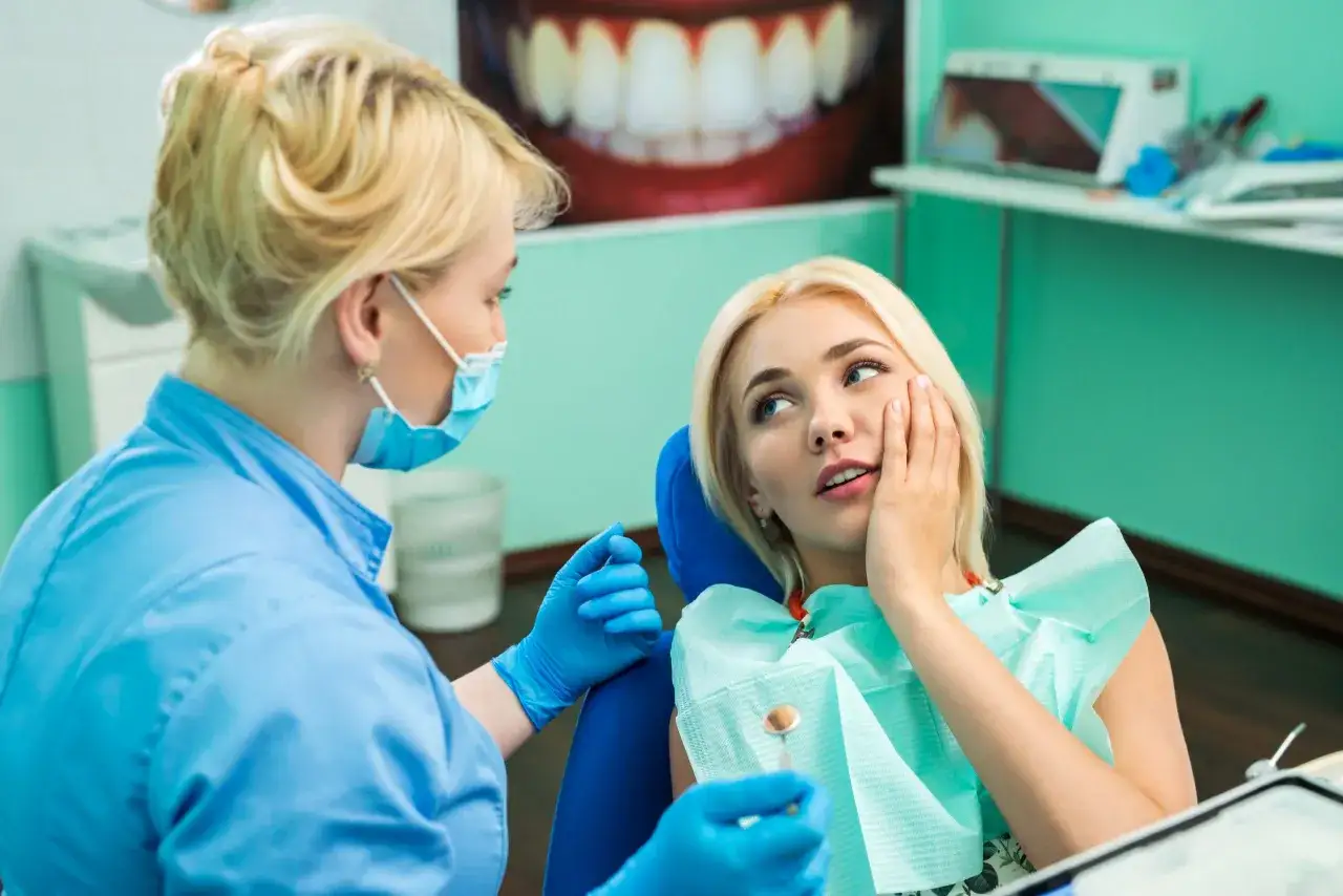 Dentystka w masce rozmawia z pacjentką, która trzyma się za policzek. Prawdopodobnie omawiają antybiotyki w stomatologii.