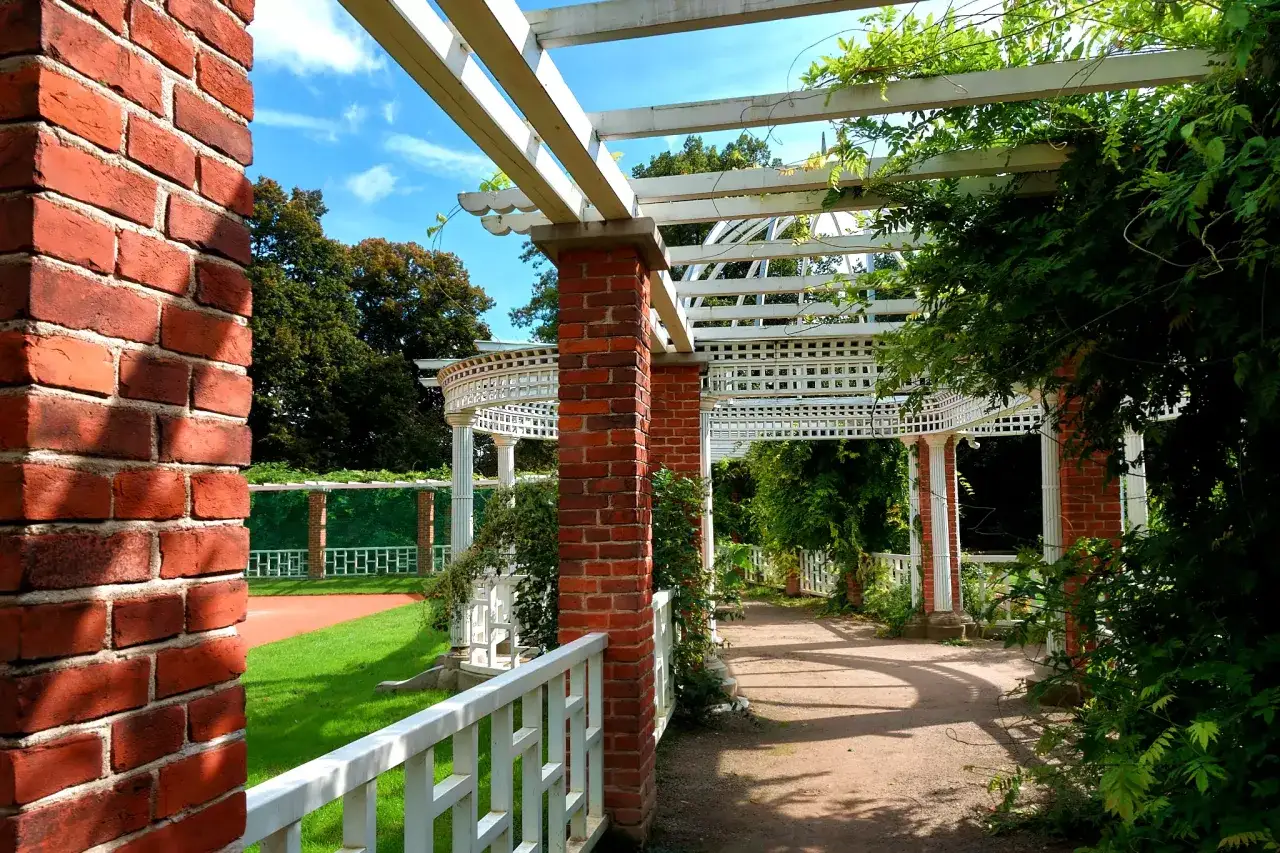Pergola z ceglanymi słupami i białymi kratownicami, otoczona zielenią. W oddali trawnik i drzewa. Jedna z urokliwych atrakcji Łańcuta.