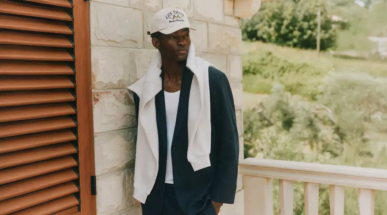Mężczyzna w bejsbolówce i marynarce, z bluzą zarzuconą na ramiona, prezentuje miejski styl streetwear.