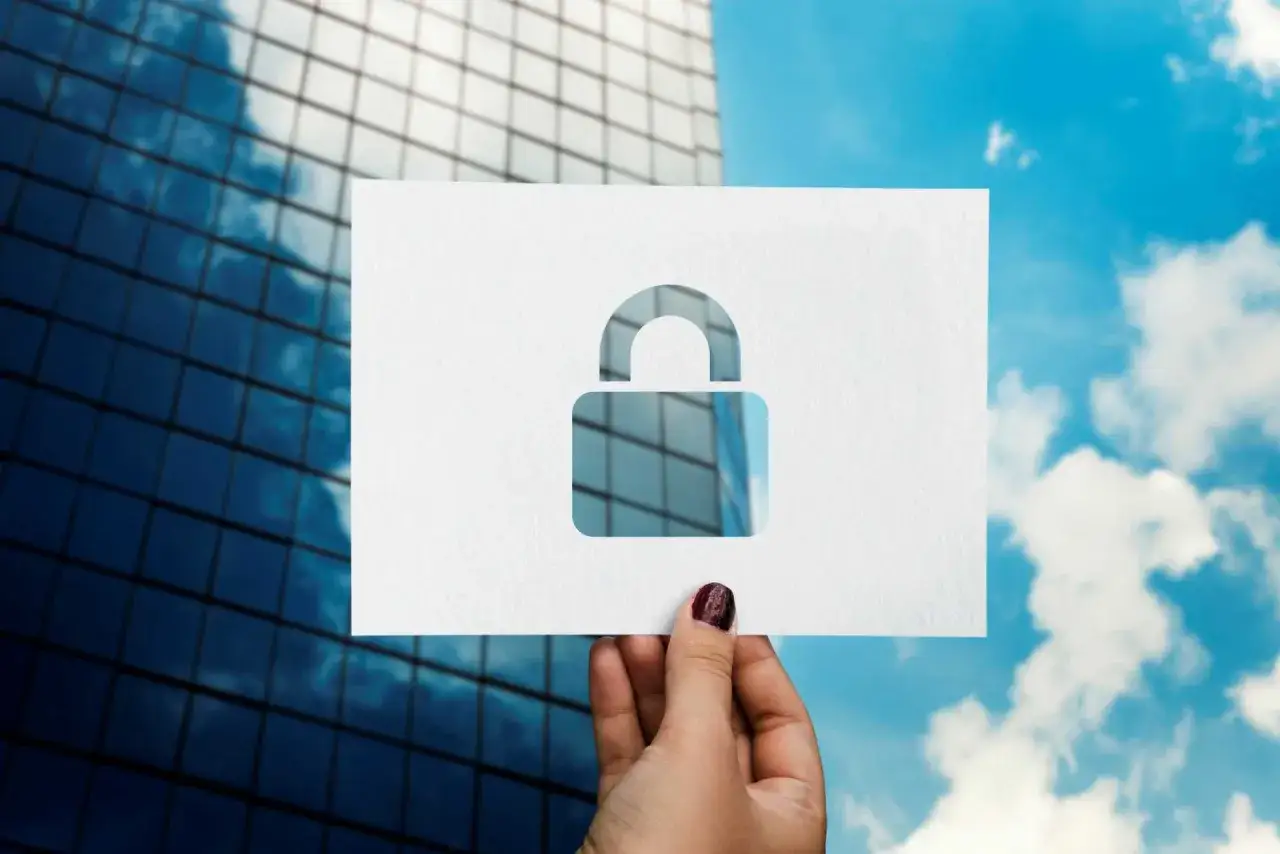 A hand holds a paper cutout of a padlock, symbolizing secure government data storage, against a backdrop of a modern skyscraper and a cloudy sky.