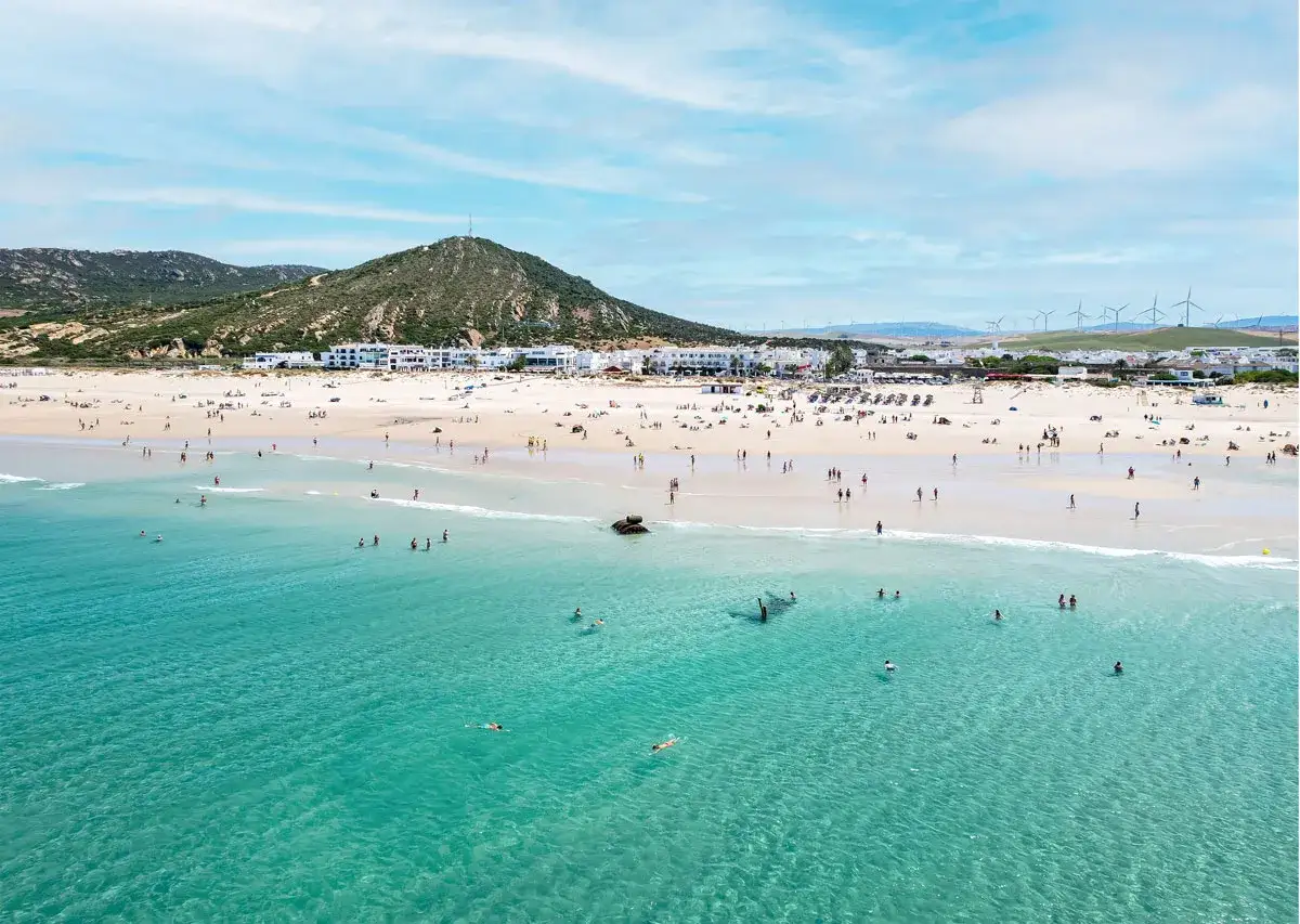 Playa de Zahara de los Atunes con gente bañándose y disfrutando del sol, con el pueblo blanco al fondo y molinos de viento en la colina.