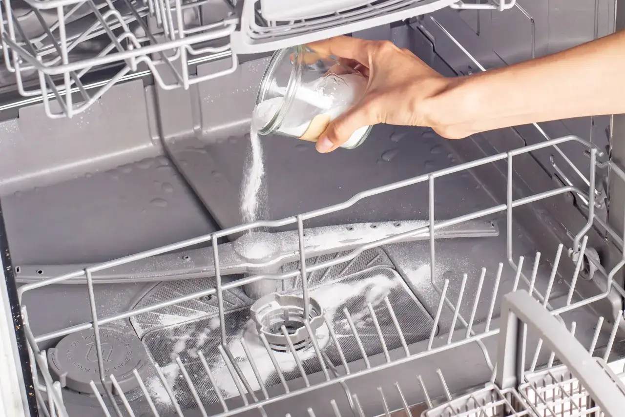 A hand pours a white powder into a dishwasher, showing how to clean a dishwasher naturally with baking soda.