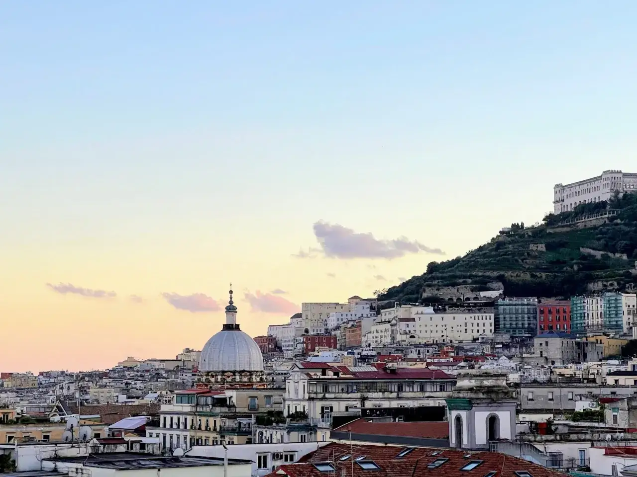 Naples cityscape at sunset. A dome and buildings climb a hill, with a grand palace at the summit. Perfect for a 2 days in Naples adventure.