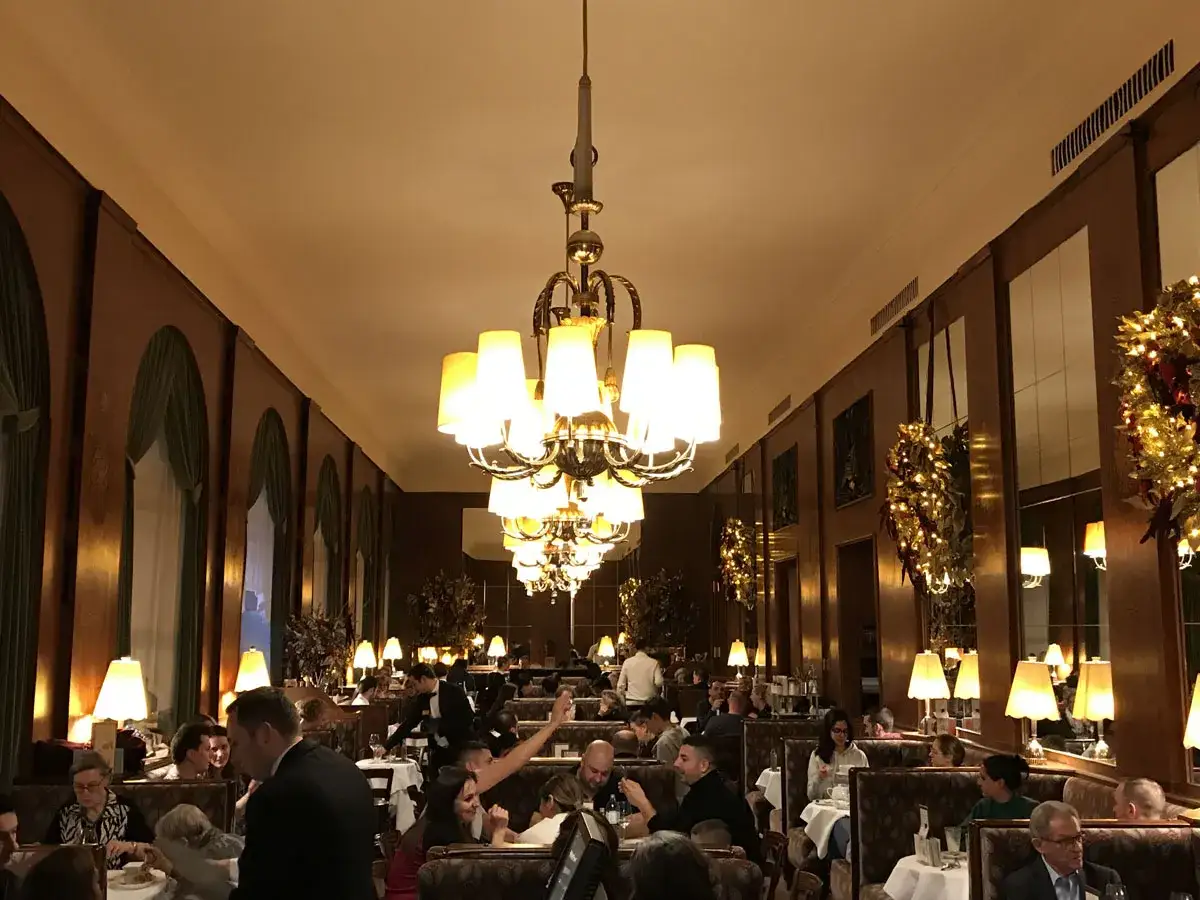 Interno elegante di uno dei caff&egrave; storici di Vienna, con tavoli apparecchiati, clienti e lampadari imponenti.
