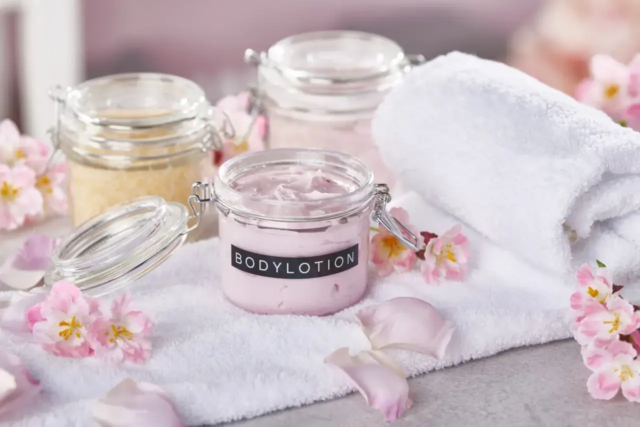 Zutaten für selbstgemachte Lotion in Gläsern, umgeben von zarten Blüten und einem weichen Handtuch.