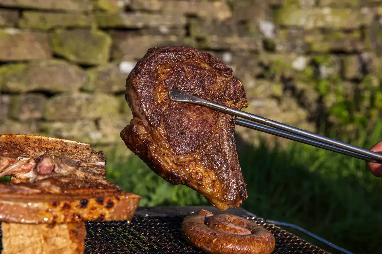Un beau morceau de bœuf des Highlands, grillé à la perfection, est soulevé par des pinces. Une saucisse enroulée cuit à côté.