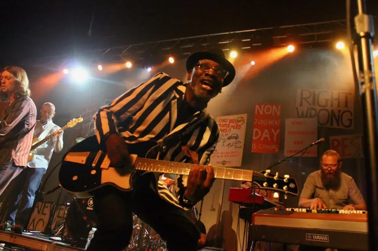 Bandmitglieder von The Specials spielen auf der Bühne. Ein Gitarrist mit Hut und gestreiftem Sakko singt.