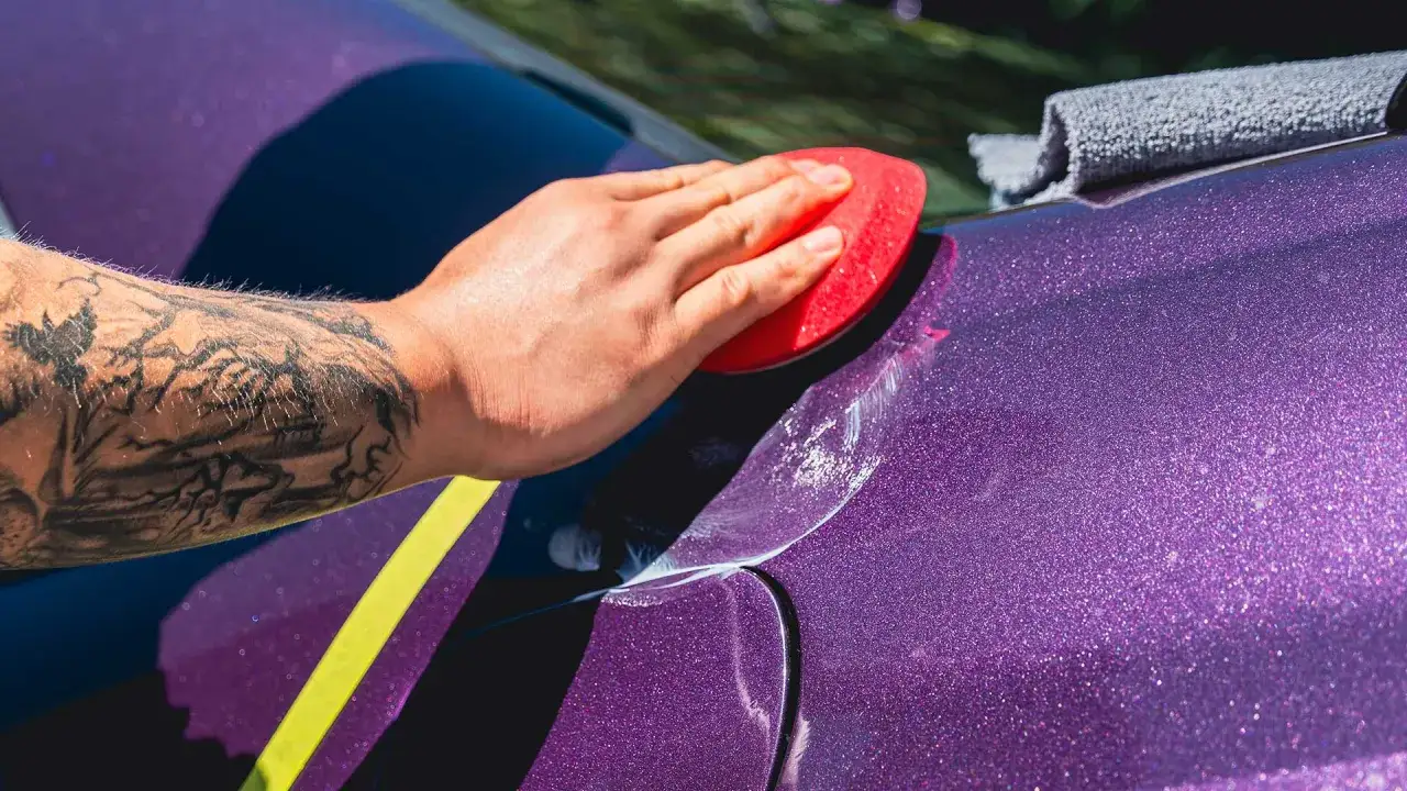 Applying multiple coats of wax to a sparkling purple car. A hand with a mountain tattoo holds a red applicator.