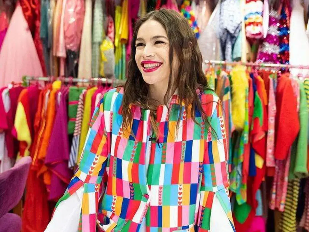 Cosima Ruiz de la Prada, sonriendo, luce una chaqueta colorida con estampado de cuadros y corazones, rodeada de ropa vibrante.