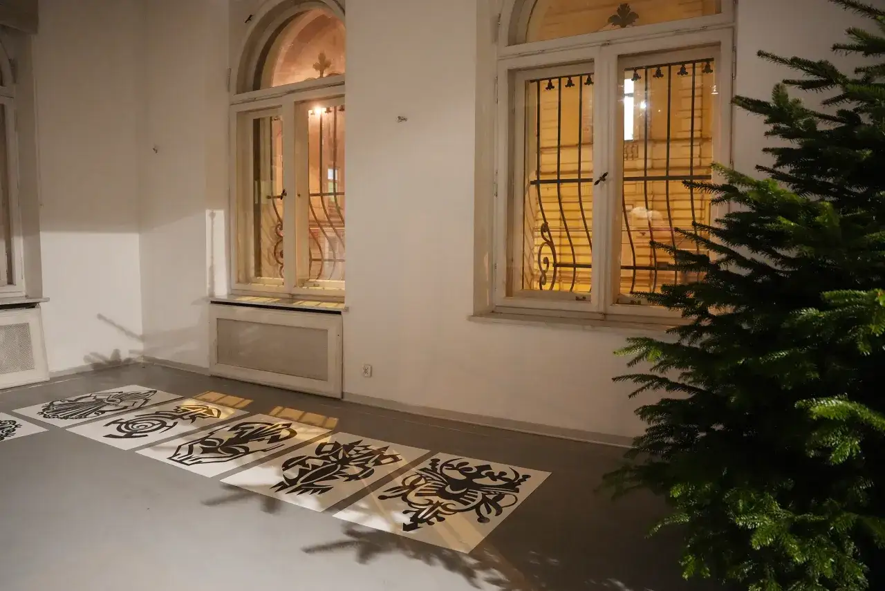 W Muzeum Sztuki Współczesnej w Łodzi, na szarym podłożu, leżą czarno-białe grafiki. W tle okna z kratami i choinka.
