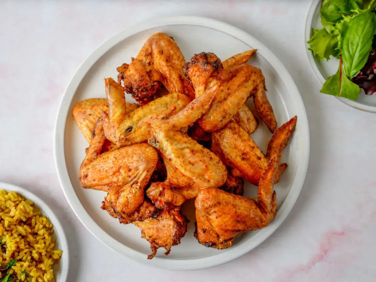 Un plato blanco lleno de alitas de pollo al horno, doradas y crujientes, listas para disfrutar. Acompañadas de arroz amarillo y una ensalada fresca.