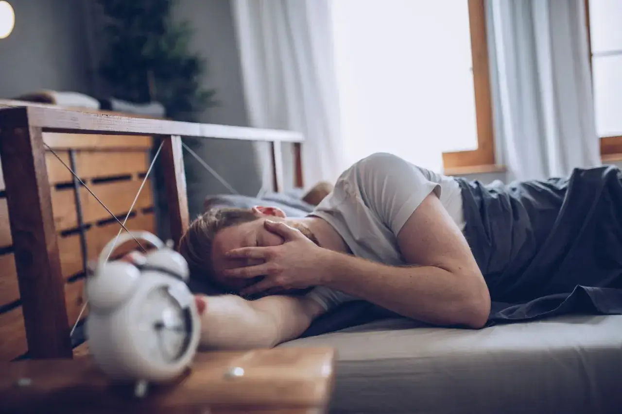 A man in bed, covering his face, experiencing paradoxical insomnia. An alarm clock sits nearby, a symbol of the struggle to rest.