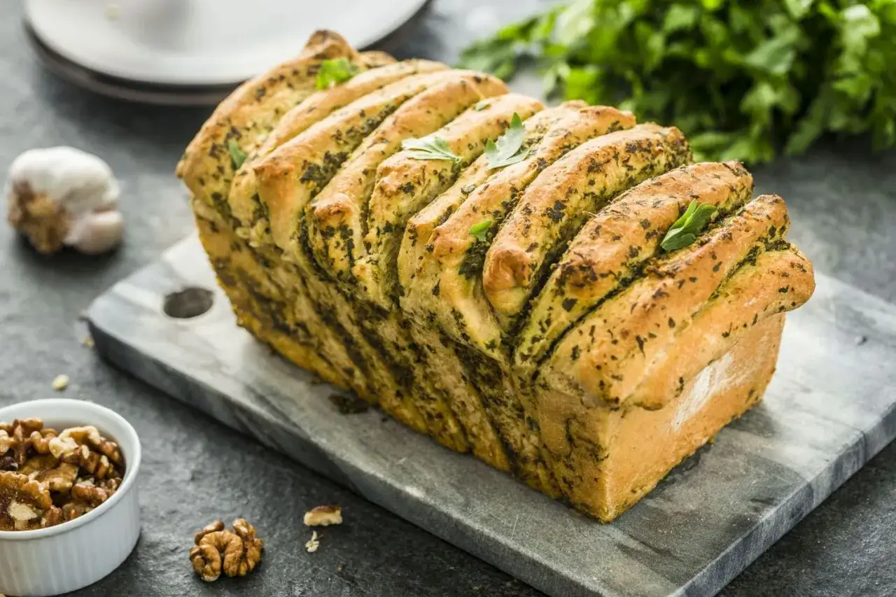 Pyszny chlebek rwany z pesto, udekorowany listkami bazylii, prezentuje się apetycznie na kamiennej desce. Obok czosnek i orzechy włoskie.