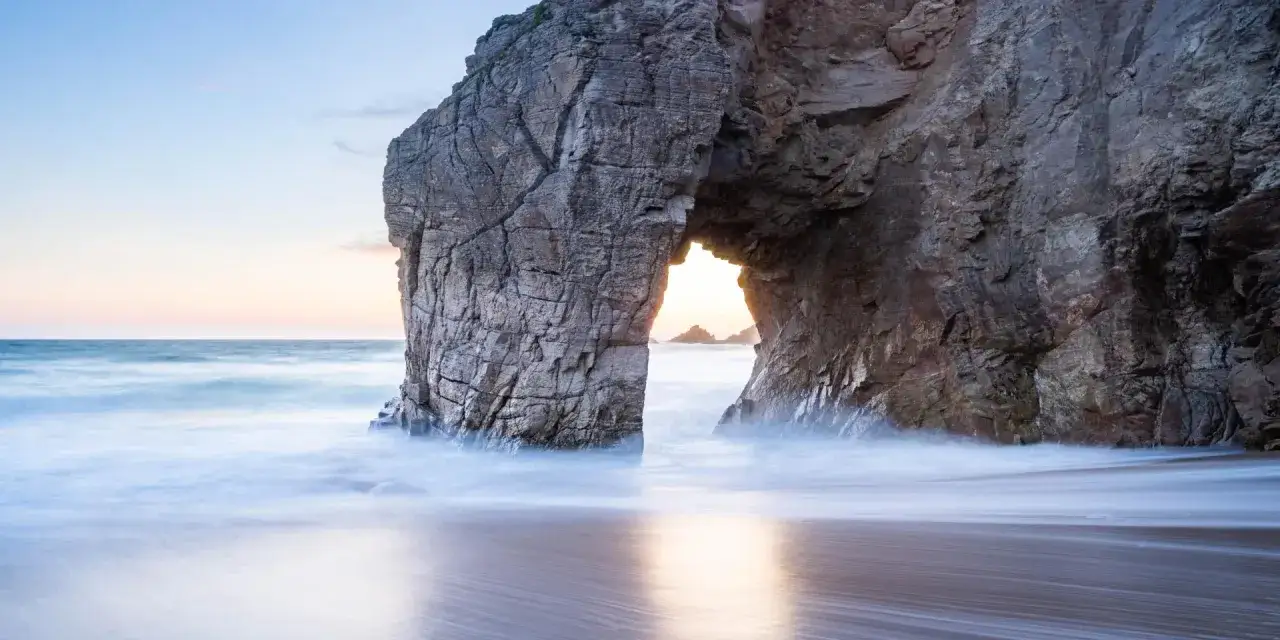 Une arche rocheuse spectaculaire sur la côte, baignée par la lumière douce du crépuscule. L'eau de mer, rendue soyeuse par une longue exposition, caresse le sable. Un aperçu des plus beaux endroits de Quiberon.
