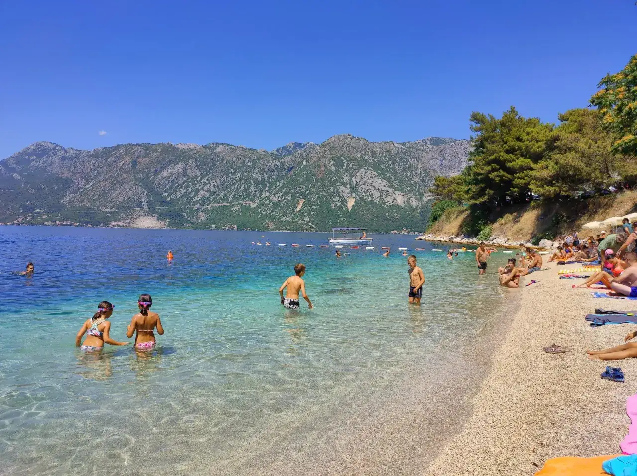 Słoneczny dzień na **Perast beach**. Ludzie kąpią się w krystalicznie czystej wodzie, podczas gdy w tle widać góry i łódź.