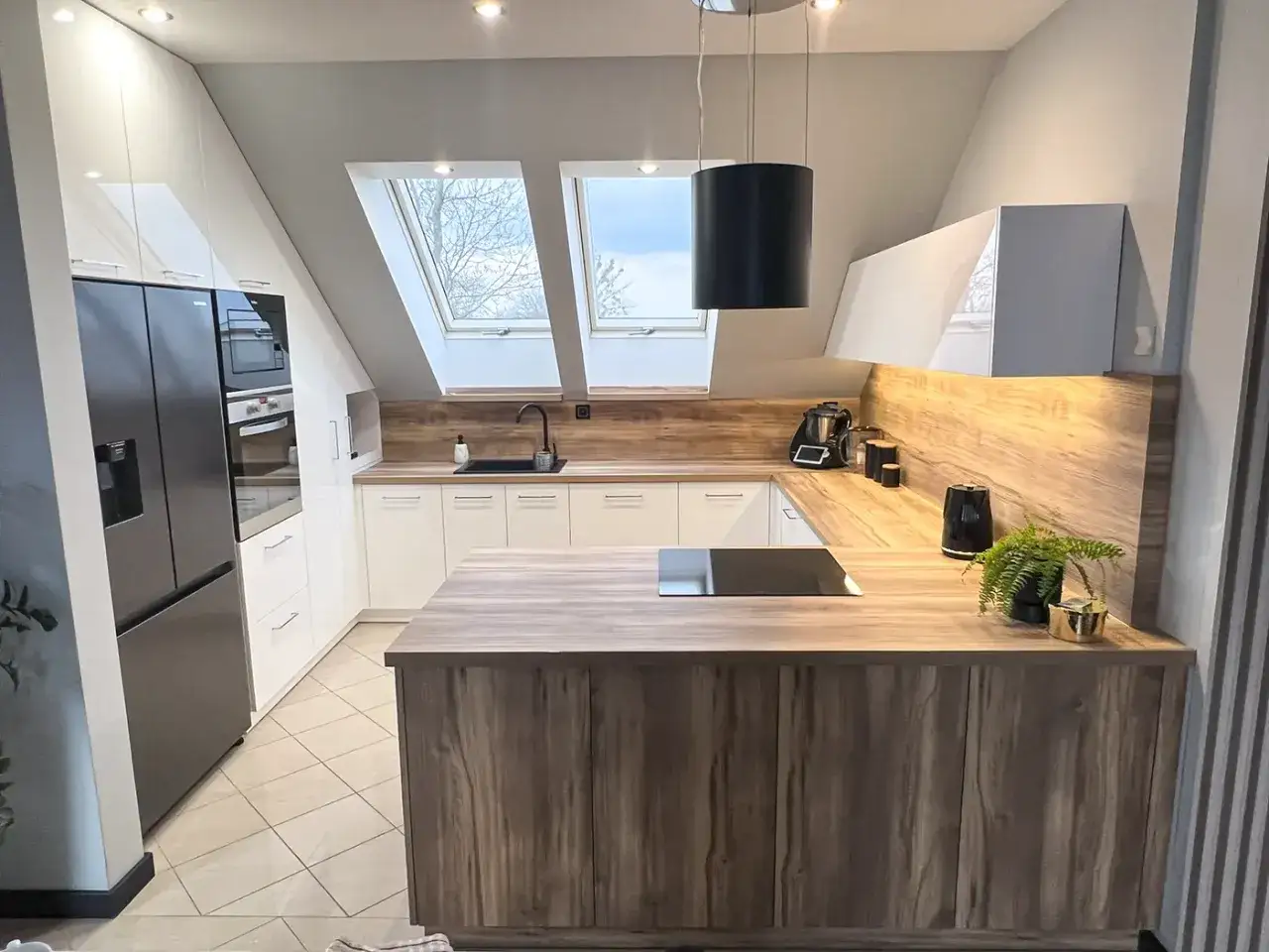 Nowoczesna kuchnia ze skosem pod skosami dachu. Białe szafki, drewniany blat i wyspa. Lodówka, piekarnik, płyta indukcyjna.