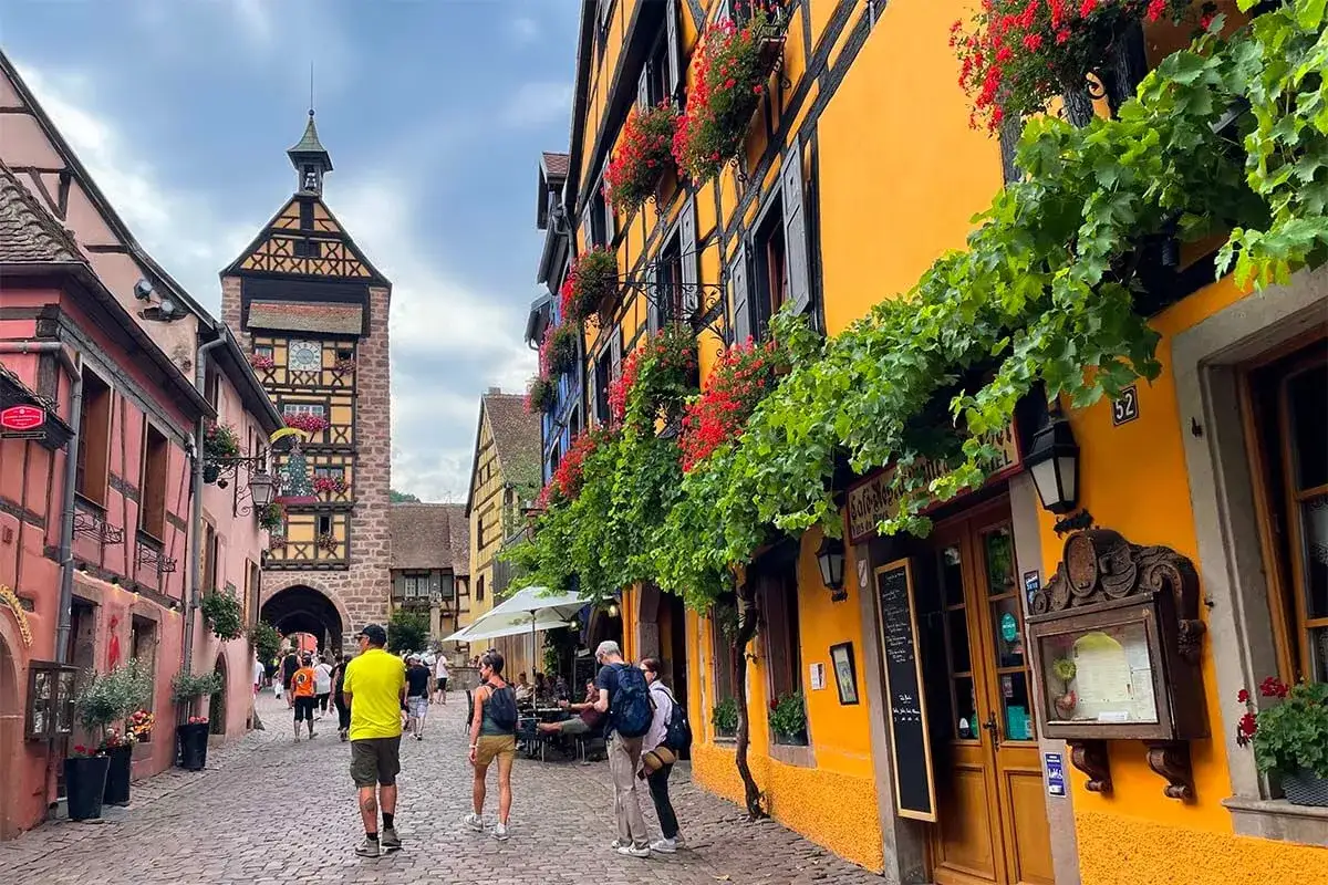 Cobblestone street in a charming Alsace town, with colorful half-timbered buildings adorned with flowers. A clock tower stands tall in the distance.