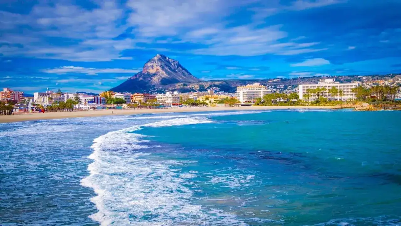 Olas turquesas rompen en la arena dorada de la playa del Arenal Javea, con edificios y el imponente Montgó de fondo.