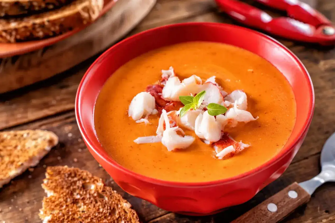 Un bol rouge vif de bisque de homard, garni de morceaux de homard et de basilic. Des toasts grillés et un casse-noix de homard rouge sont visibles en arrière-plan.