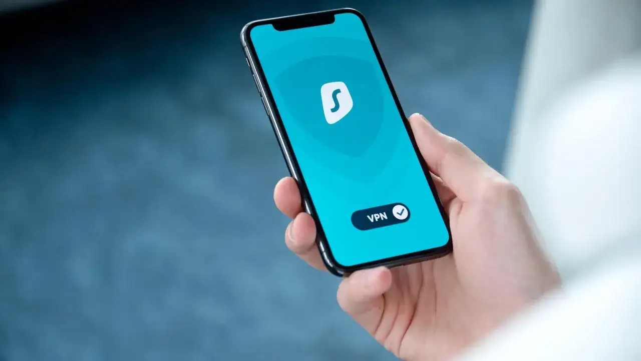 Dłoń trzyma smartfon z włączonym vpn na telefon. Aplikacja ma niebieskie tło i białe logo.