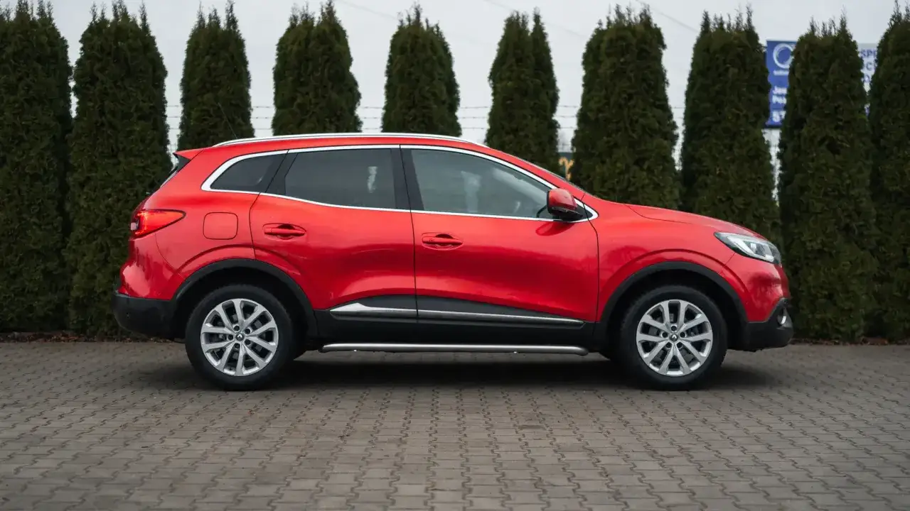 Czerwony Renault Kadjar, jeden z popularnych renault modele, zaparkowany na tle zielonych żywotników.