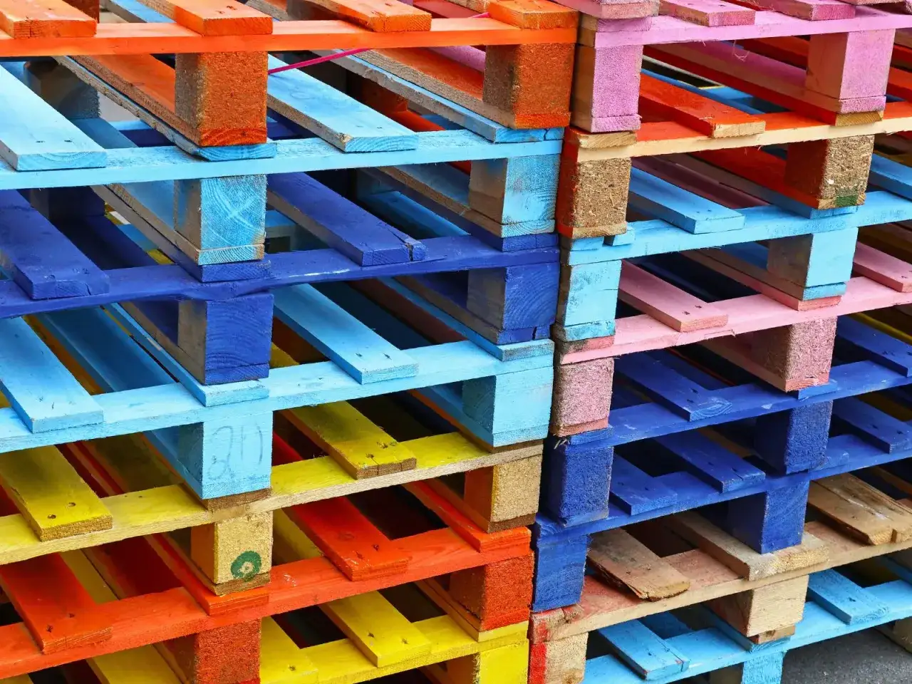 Palets apilados en vibrantes colores: azul, rosa, naranja y amarillo. Una gu&iacute;a visual para como pintar un palet y darle vida a tus proyectos.