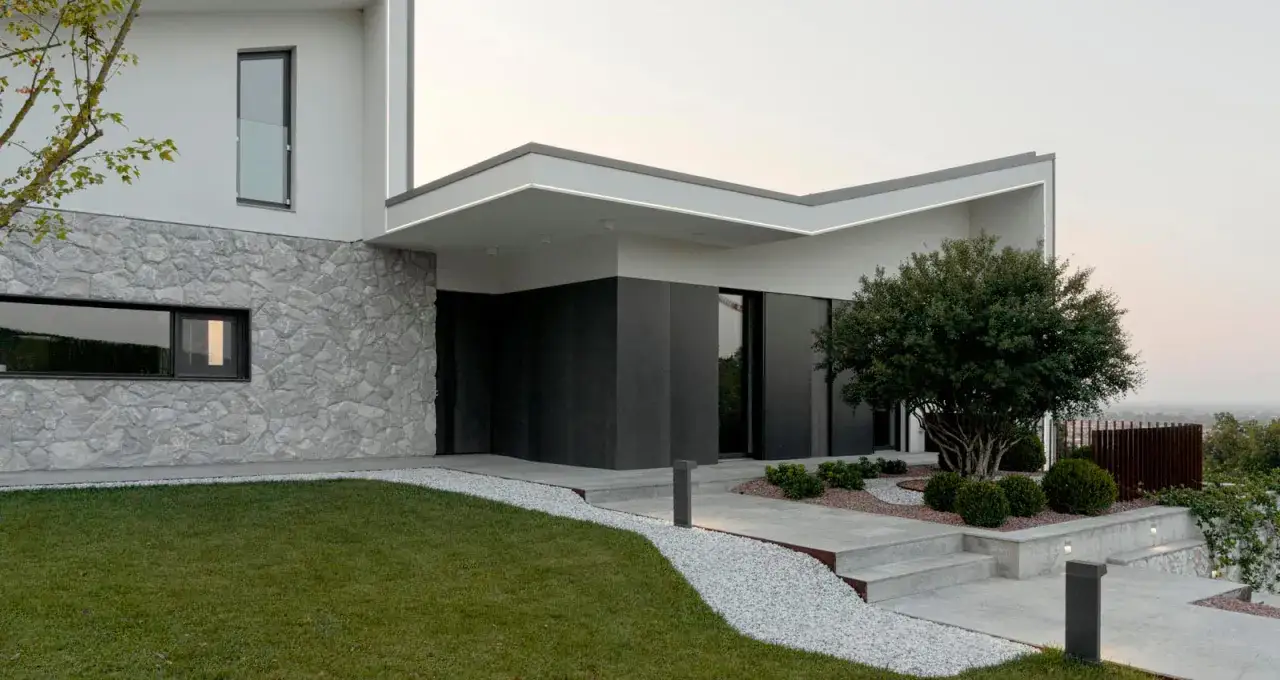 Moderna villa con facciate in pietra grezza e dettagli in cemento scuro. Un vialetto di ghiaia bianca conduce all'ingresso.