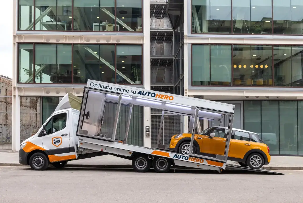 Un furgone bianco con logo AUTOhero trasporta un'auto gialla. La scritta