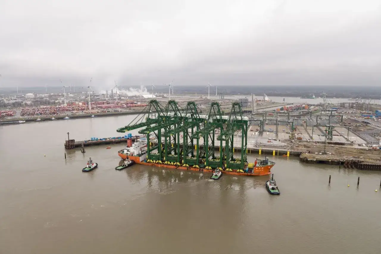 Schwere Kr&auml;ne werden im Antwerpen Port auf einem Schiff transportiert, umgeben von Schleppern und der weitl&auml;ufigen Hafenanlage.