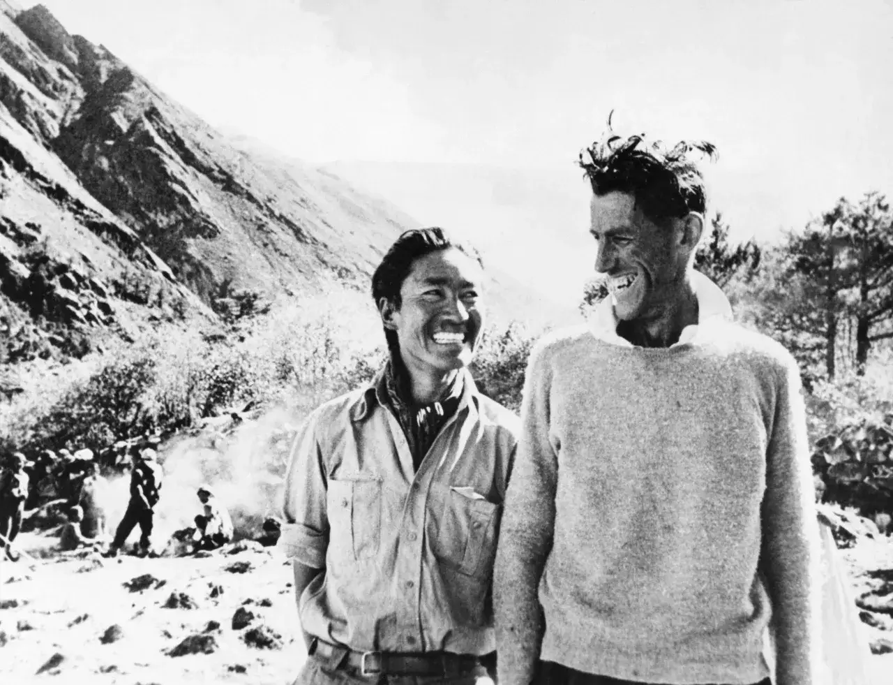 Tenzing Norgay i Edmund Hillary śmieją się po pierwszym zdobyciu Mount Everest. W tle widać zbocza g&oacute;ry i innych członk&oacute;w wyprawy.