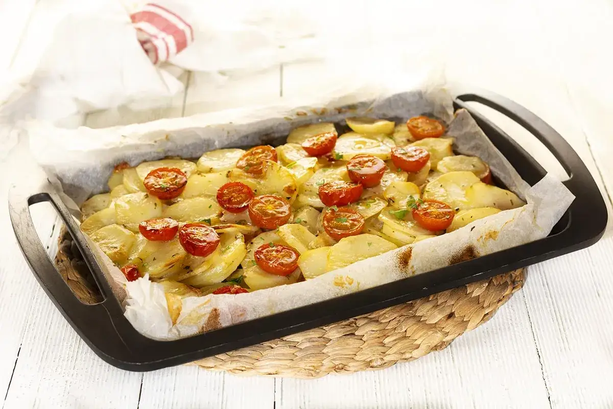 Delicioso calabacín al horno con patatas y tomates cherry, servido en una bandeja sobre una esterilla de mimbre.