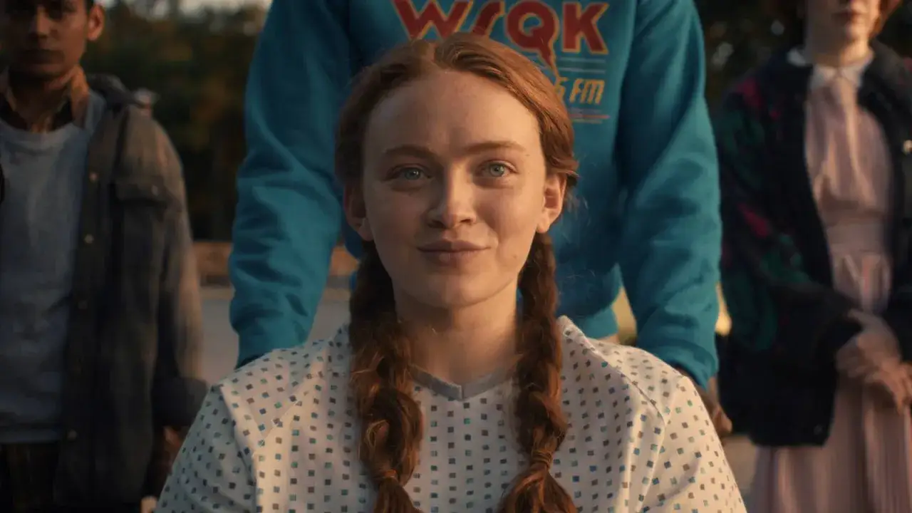 Maxine Mayfield, avec ses tresses rousses, sourit doucement, entourée de ses amis. Elle porte une blouse à petits carreaux bleus.