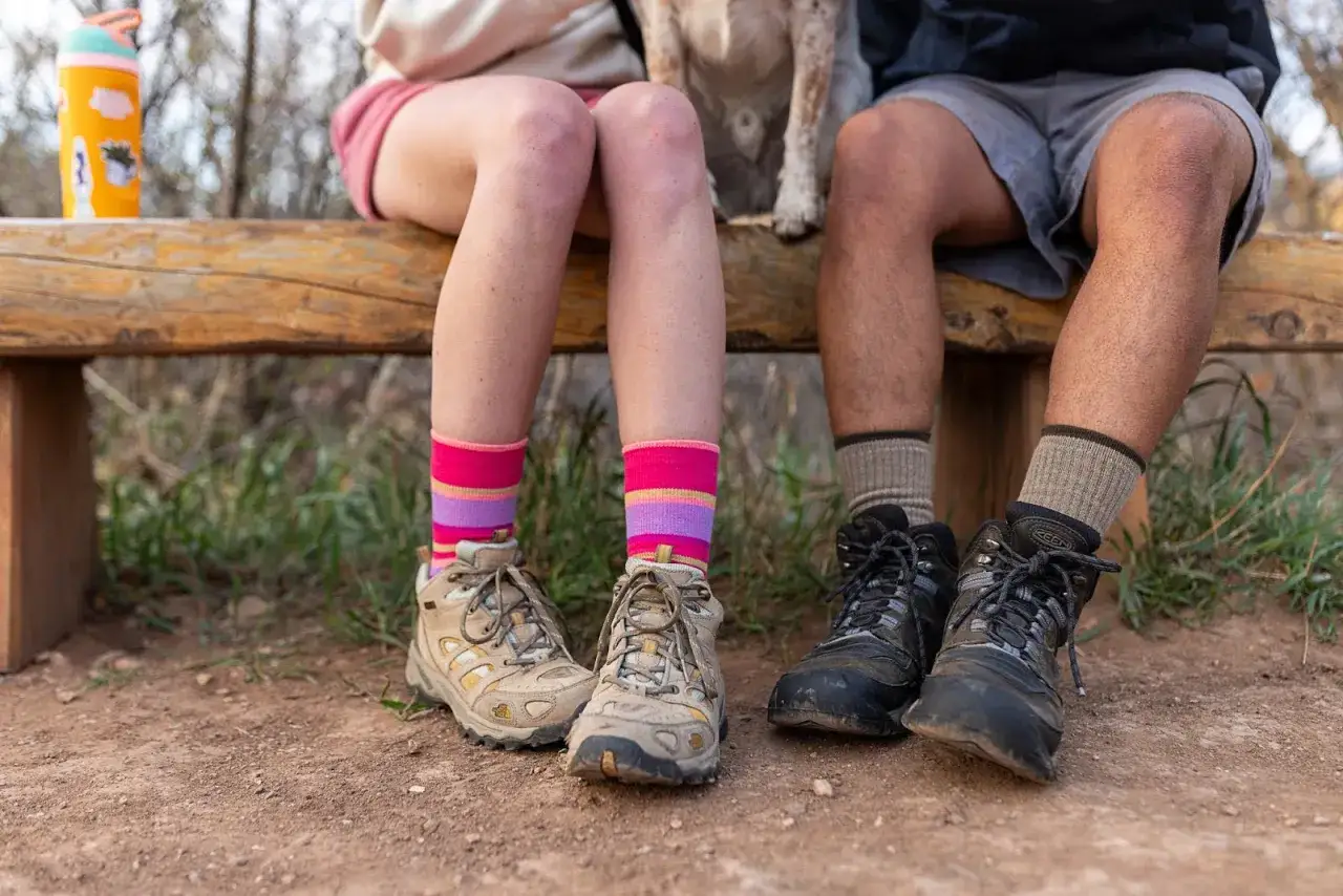 Two people sit on a bench, one wearing colorful striped socks and hiking boots, the other in plain socks and boots. Is wearing two pairs of socks bad?