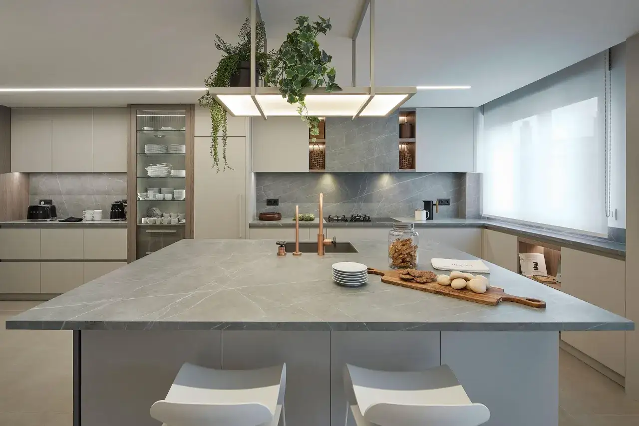 Moderna cocina con isla central de m&aacute;rmol gris. Encimeras a juego, armarios minimalistas y grifer&iacute;a de cobre. Plantas colgantes a&ntilde;aden un toque verde.