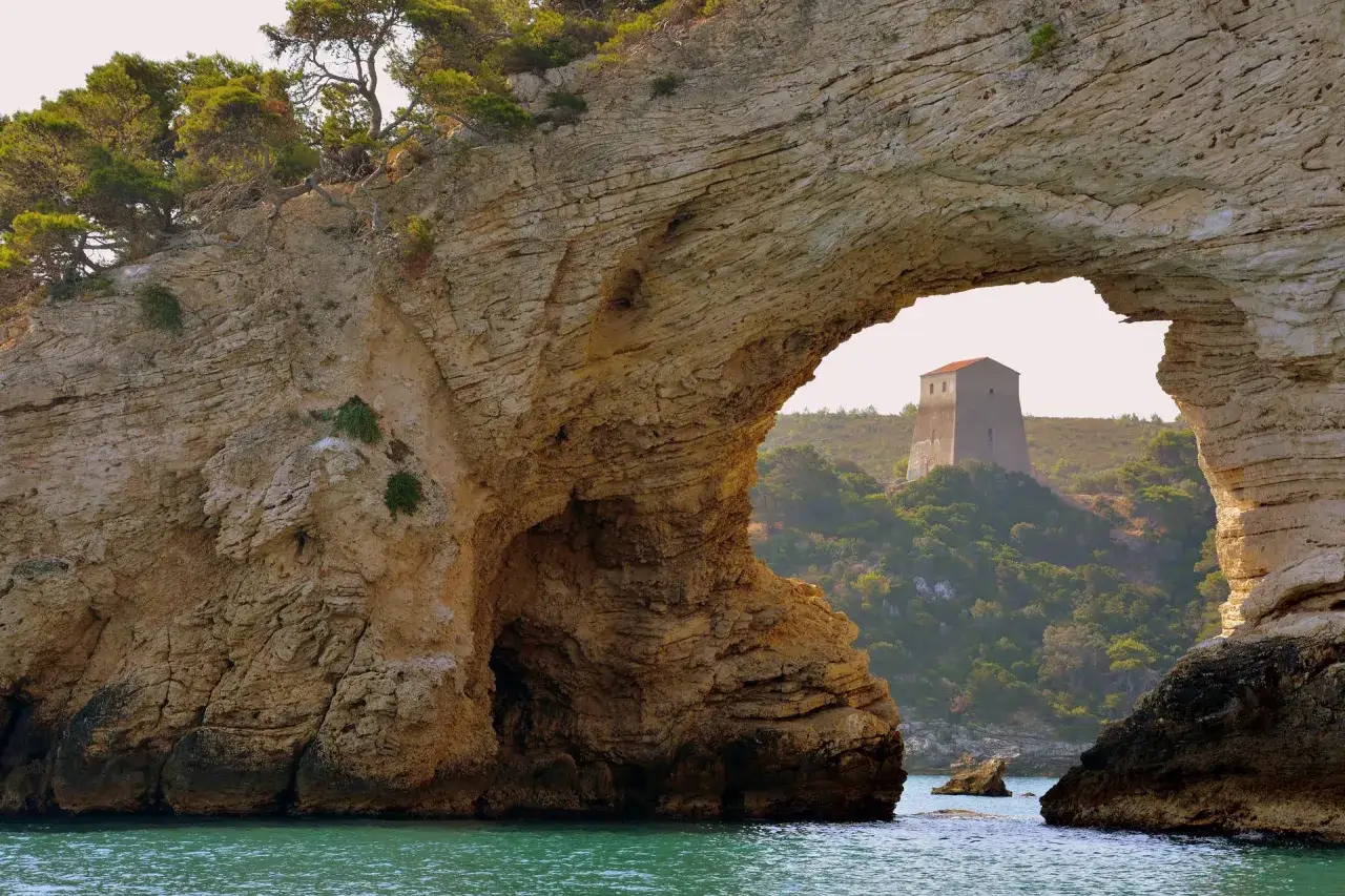 Arco naturale roccioso che incornicia una torre antica su una collina verdeggiante. Baia San Felice Vieste, Puglia.