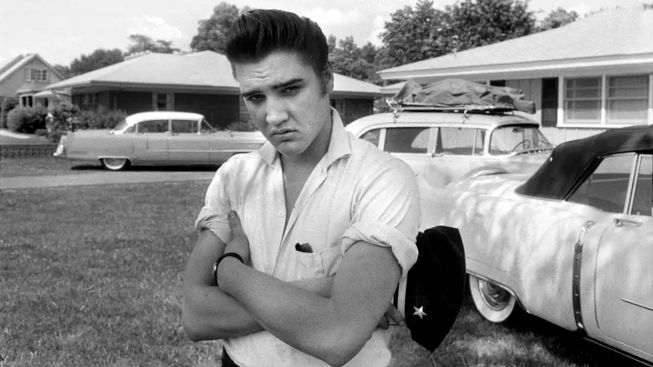 Elvis Presley, mit verschränkten Armen und einem ernsten Blick, steht vor seinen Autos. Sein Vermögen, der Elvis Presley net worth, war schon damals beeindruckend.