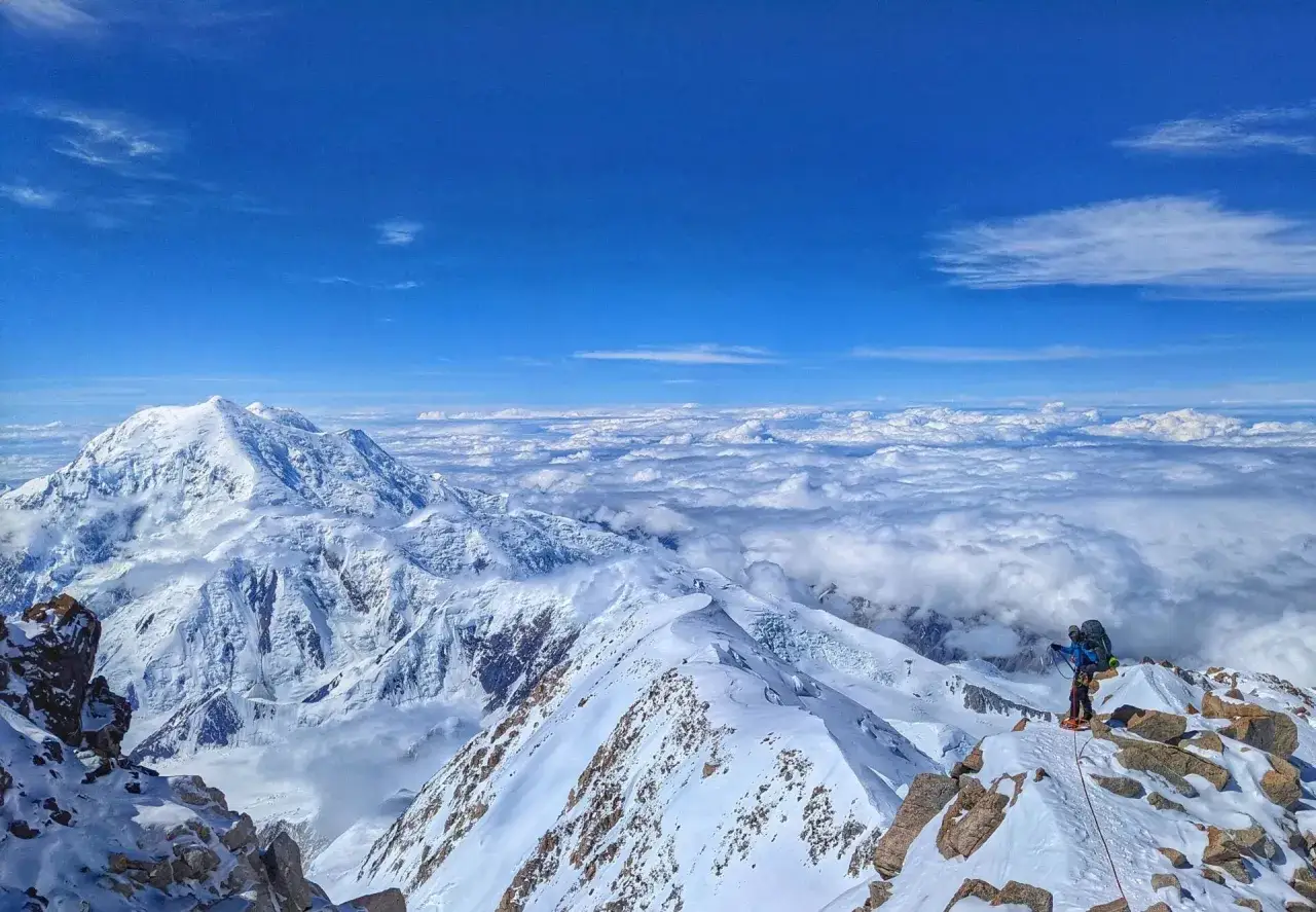Alpinista na szczycie najwyższego szczytu Ameryki Północnej, Denali, pod błękitnym niebem, nad morzem chmur.