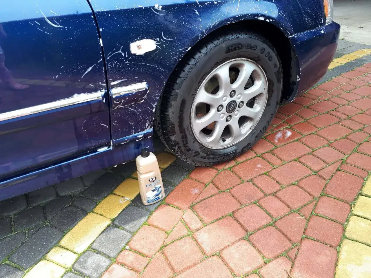 Butelka wosku do auta stoi obok umytego, niebieskiego samochodu. Woskowanie auta daje połysk i ochronę.