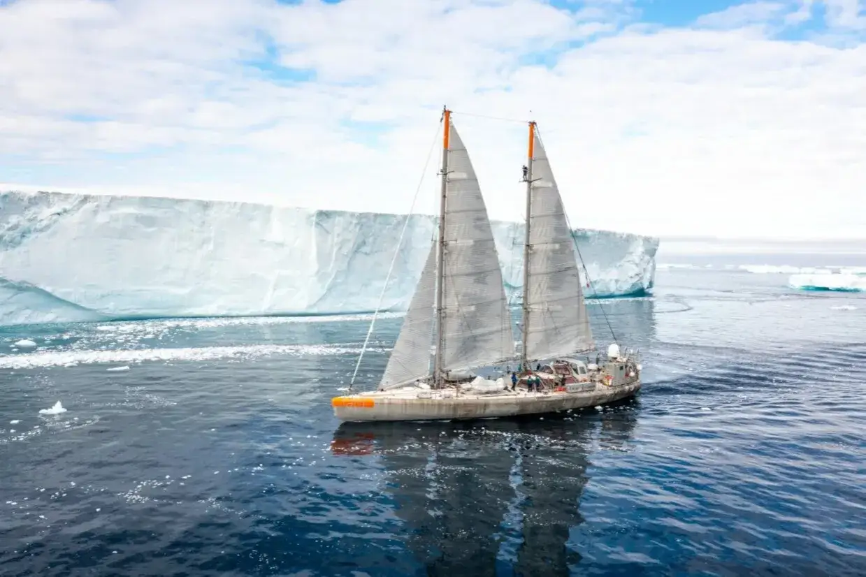 Le Tara bateau navigue près d'un immense iceberg sous un ciel bleu et nuageux.
