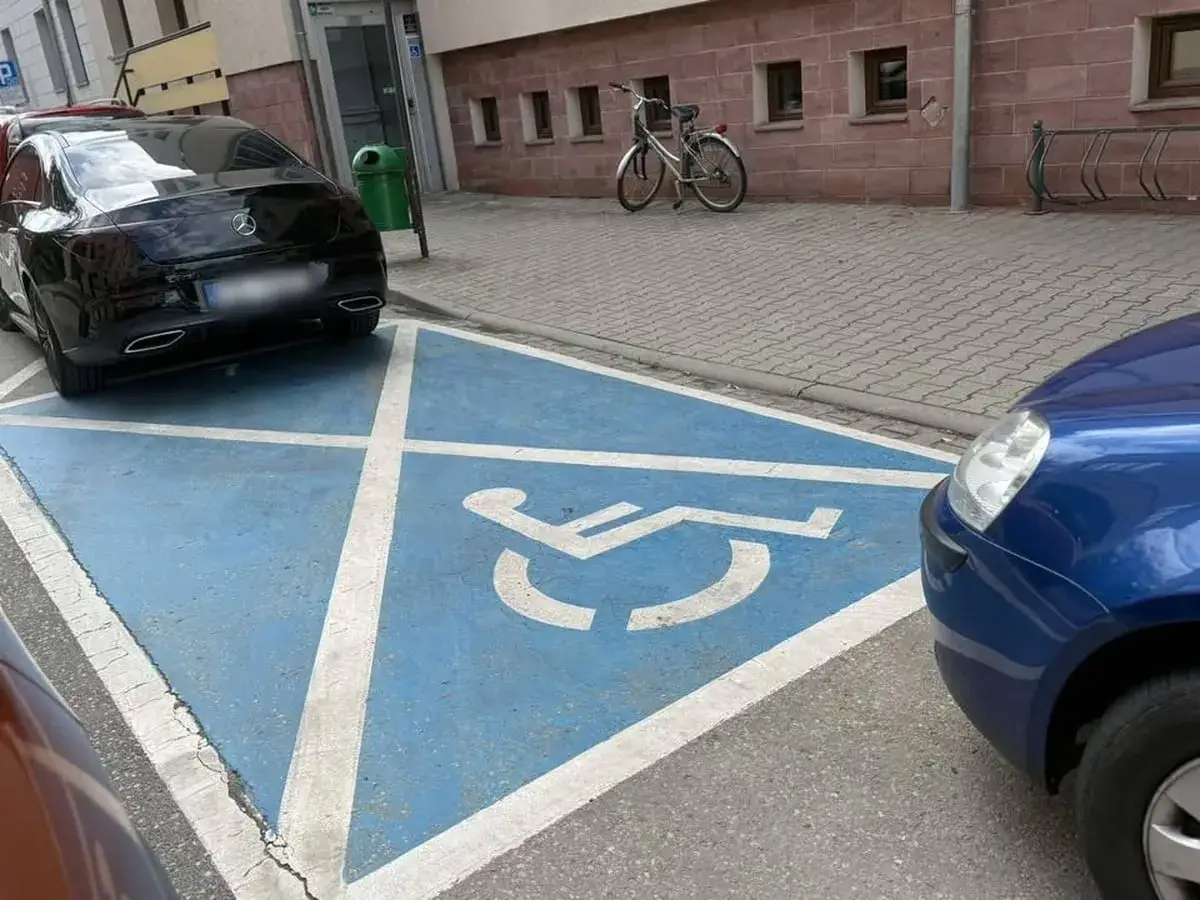 Czarny Mercedes zaparkowany na niebieskim polu z symbolem wózka inwalidzkiego. Ktoś pewnie zaraz dostanie mandat za parkowanie na miejscu dla niepełnosprawnych.