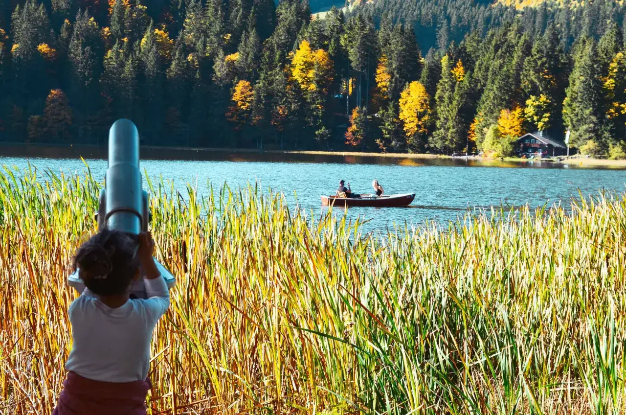 Ein Kind blickt durch ein Fernglas auf einen ruhigen See, umgeben von bunten Herbstw&auml;ldern. Ein Boot mit zwei Personen gleitet &uuml;ber das Wasser. Ein malerischer Anblick am M&uuml;nchner See.