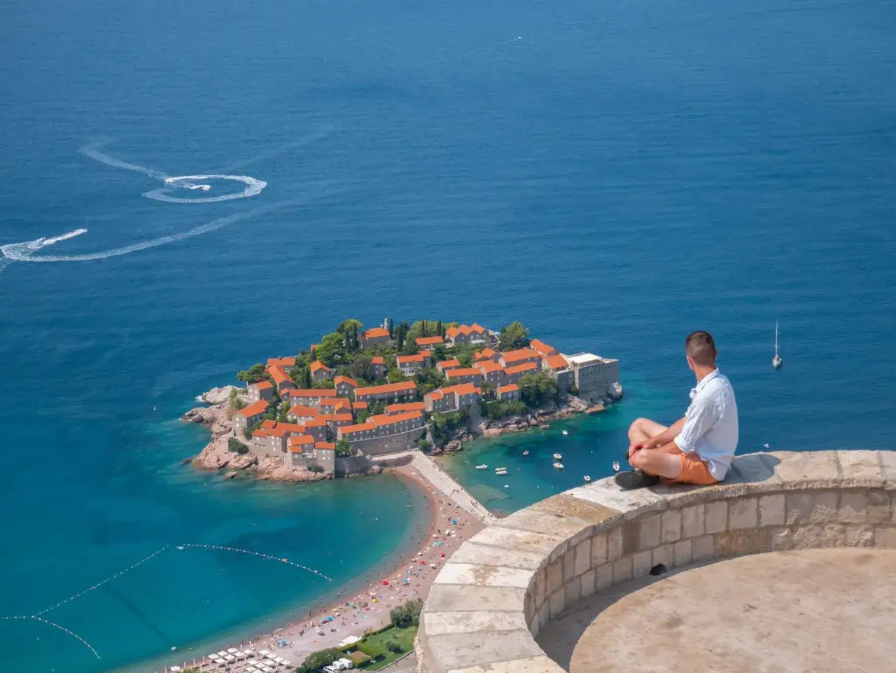 Mężczyzna siedzi na murze, podziwiając wyspę Sveti Stefan. Czy Czarnog&oacute;ra jest bezpieczna? Tak, wygląda na spokojną i malowniczą.